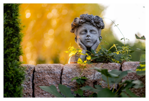 Kleine Büste eines blütengekrönten Knaben auf einer Mauer. Von hinten links scheint die Abendsonne warm und gelb.
