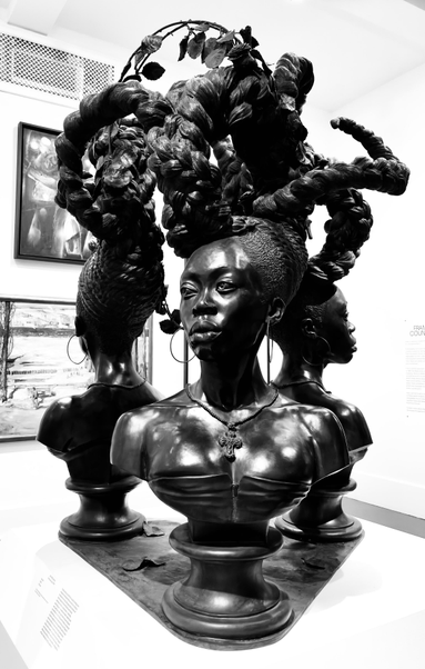 Black and white photo of Kehinde Wiley’s bronze sculpture BOUND (2014), three busts of Black women whose hair rises and intertwines above their heads.

From the Brooklyn Museum’s description: “In Bound, the artist's first sculptural depiction of women, Kehinde Wiley constructs a tribute to Black femininity. Occupying the corners of the triangular base, the busts are nearly identical in their countenance—a steady, piercing gaze—as well as in their adornment and, perhaps, faith, given the crosses draped around their necks. They are further connected by their hair, which extends from each figure's head in a tightly braided updo that widens and loosens into an entwined flourish above their heads. Embellished with leaves, flowers, and birds, their crowns are given as much space and respect as the subjects themselves. Here, Wiley honors the beauty, function, and politics of hair in the lives of Black women.”