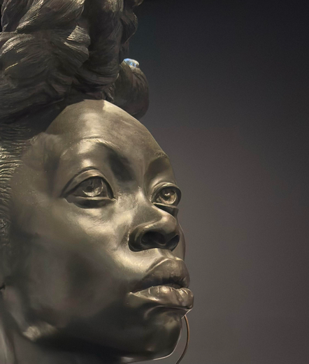 A closeup of Kehinde Wiley’s sculpture BOUND, focusing on the face of one of the bronze busts against a dark background. The Black woman’s face is in three-quarter view with her gaze directed towards the upper right frame. Her expression is somber, steely, confident, determined. Some intricate details of her hair are visible, as is part of a large hoop earring hanging from her unseen left ear.