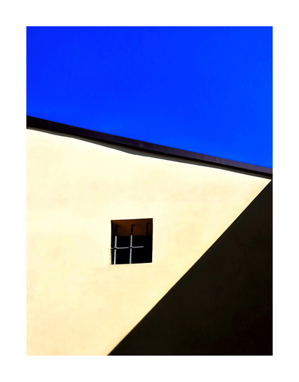 This is a photograph of the upper corner of a minimalist, modern building. It is a large, flat, beige wall with a small, square, black-framed window near the centre. The background is a clear blue sky.