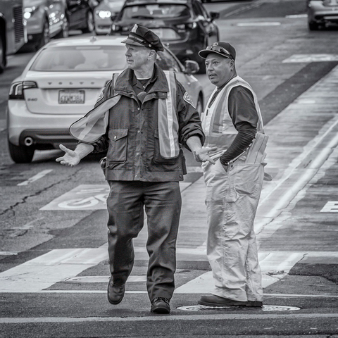 Traffic cop and road worker stand in an intersection in San Francisco. The cop is holding his arms out with an expression of disbelief on his face as both men look at something to left.