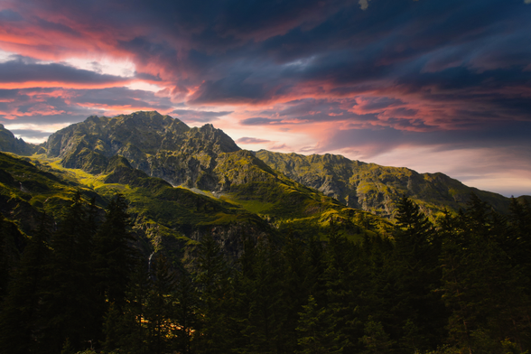 A vibrant landscape featuring rugged mountains under a colorful sky at sunset, with lush greenery in the foreground and dark trees framing the scene.