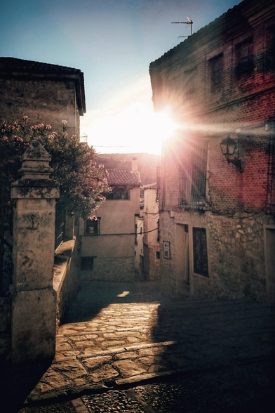 A narrow, sunlit street in a historic Spanish town. The sun flares brightly between two stone buildings, casting long shadows on the cobblestone path. A flowering oleander bush sits to the left, adding a touch of colour. The warm light and old buildings create a sense of timelessness and peace.