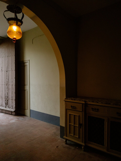 Dimly lit hallway with an arched doorway, an antique wooden sideboard, and a warm amber hanging lamp casting a soft glow.