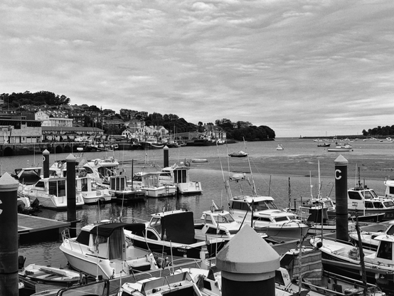 Una fotografía en blanco y negro de un puerto con numerosos barcos amarrados. En primer plano, se ven de cerca varios barcos de recreo y pesca, de tamaño pequeño a mediano, algunos de ellos atados a postes oscuros que tienen una letra "C" blanca. El agua del puerto parece tranquila, y en la parte media de la imagen, la marea baja ha dejado al descubierto áreas de lodo y arena, con algunos barcos adicionales anclados más lejos. Al fondo, se distingue un pueblo costero con casas y edificios escalonados sobre una colina densamente arbolada. El cielo está cubierto de nubes, lo que genera una luz suave y difusa sobre toda la escena.