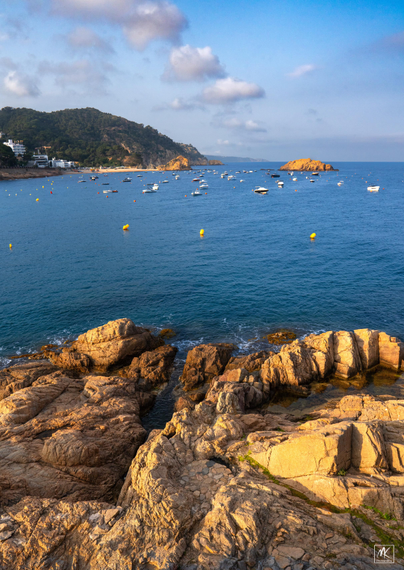 Color photo taken late in the afternoon with a rocky shoreline in the foreground, a small bay with moored boats in the middle ground, and a hilly coastline stretching off into the background distance. 