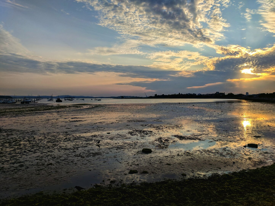 A tranquil sunset scene over a wide expanse of exposed mudflats and shallow water. The tide is out, revealing a textured, reflective surface with small rocks and pools of water mirroring the dramatic sky. In the distance, a calm body of water contains several small boats moored along the left side. The horizon is a mix of a silhouetted treeline and buildings. The sun, partially hidden by clouds on the right, casts a golden and orange glow across the water and into the sky. The clouds above are wispy and streaky, painted with soft hues of pink, yellow, and blue.