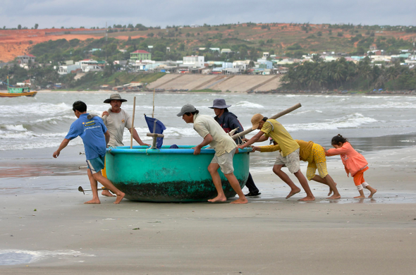 das foto zeigt eine gruppe von menschen aus einem vietnamesischen küstenort, die ein türkisfarbenes boot über einen sandstrand zum meer ziehen. es sind erwachsene und kinder beteiligt, die zusammenarbeiten, um das boot zu bewegen. im hintergrund ist ein dorf mit häusern und palmen zu sehen sowie ein meer mit kleinen wellen. der himmel ist bewölkt.
das bild vermittelt einen eindruck von gemeinschaftlicher arbeit und dem täglichen leben in einem küstenort. die farben sind gedämpft und die stimmung eher ruhig.