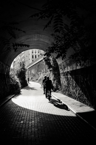 Two individuals ride bicycles through a shadowed tunnel. The tunnel itself is constructed of stone, with foliage growing along the sides, and a building can be seen in the distance beyond the tunnel's opening. A lone figure walks ahead in the distance on the other side of the tunnel. 