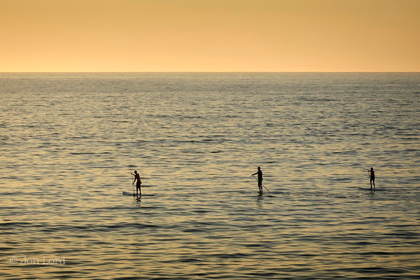 This is a colour photo of three people paddle boarding on the sea at sunset.
Stretching upwards three quarters in the frame is a calm pacific ocean with only small wavelets disturbing the surface. The setting sun is giving the sea a slight gold tint to the blue surface. There is a crisp and defined horizon with a caramel coloured clear and cloudless sky filling the remainder of the image. A third of the way up in the frame are three paddle boarders, standing on their boards and moving from right to left. The first is below and left of centre, the middle one is below and to the right of centre, the last one is close to the right margin. There is an even spacing of a few metres between them.
The location: Redondo Beach, California (2013).