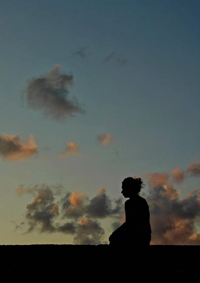 A silhouetted figure sits against the twilight sky filled with clouds tinted orange and pink from the setting sun. 