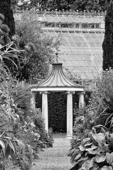 A black and white photo of a small stone gazebo with two columns and a domed roof, surrounded by lush foliage. A gravel path leads to the gazebo, with a large glasshouse visible in the background.