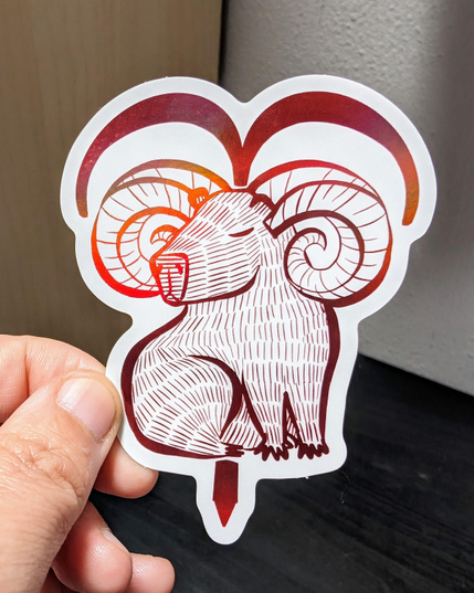 Photo of a left hand holding a diecut sticker. The sticker is a holographic drawing of a capybara Aries zodiac sign.