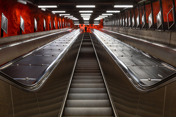 Low angle view on a dark metro elevator between uneven red walls reminding of hell in a Tunnelbana station in Stockholm. At the top of the elevator, the little black silhouette of a man who is about to go down.
