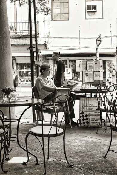 Black and white photo of an outdoor café. A person in a cap reads a newspaper at a metal table. A passerby walks by. Calm, nostalgic atmosphere.