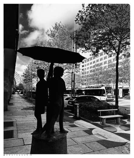 Schwarzweißfotografie: Fußweg in der Innenstadt mit der Brunnenplastik „Jugend“ von Johannes Belz, 1965 (auch bekannt als Jugendliche unter einem Regenschirm oder Regenschirmbrunnen) nur in dunklen Umrissen erkennbar, rechts parkende Autos zwischen jungen Bäumen, dahinter eine fahrende Straßenbahn und ein großes Gebäude