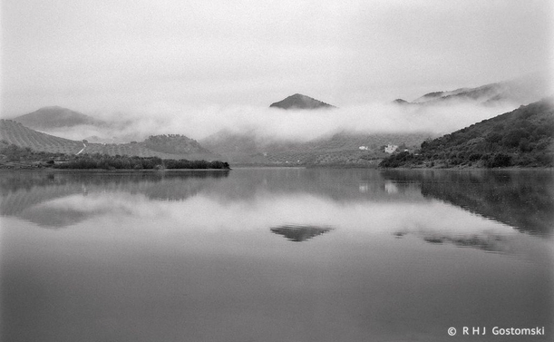 Black and white photo looking out overall a lake with low cloud and mountains in the background. The mountains and clouds reflect in the lake.