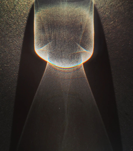 Funnel of light

Sunlight passing through a glass on a black office table. Part of the shadow turns a deeper black around the figures made from light being diffracted through water and glass.
Here a broad tunnel of light seems to hit a narrowing. On the other side the light falls to the sides and in the middle a structure reminiscent of a whirlpool can be seen passing from the tunnel through the narrow opening