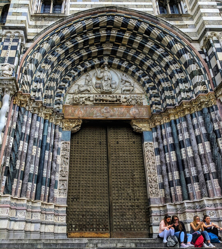 A medium shot from a low angle captures the grand entrance of the Cattedrale di San Lorenzo (Genoa Cathedral) in Italy. The large, double doors are made of dark metal with a geometric, textured pattern. They are framed by a series of concentric arches with a striking black-and-white striped pattern and detailed sculptures, characteristic of Genoese Gothic architecture. Above the doors, a large, carved stone relief depicts religious figures and animals. On the right side of the image, four young women are sitting on the steps in front of the door. The building facade above the entrance features the same black-and-white striped pattern and arched windows.