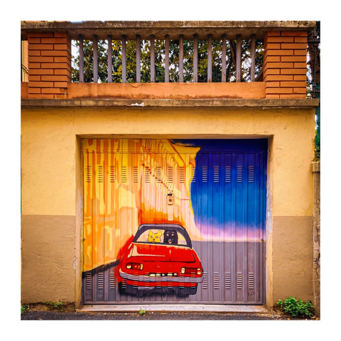 This is a vibrant photograph of a colorful mural on a yellow and beige building's garage door. The mural has a red vintage car in front of a yellow and orange building, driving towards a blue and purple gradient sky. The garage door is made of metal, and the building features a brick balcony with a railing above it. Inside the car, looking out the back window are two dogs. One beige and one black.