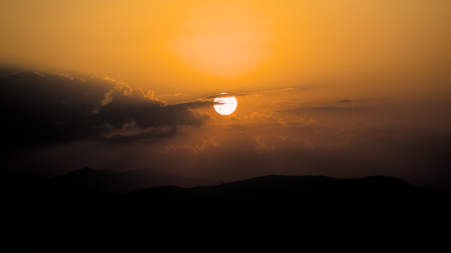 Berge im Dunkeln, darüber der runde Sonnenball, teilweise hinter Wolken. Die Stimmung geht von Schwarz bis ins Orange.