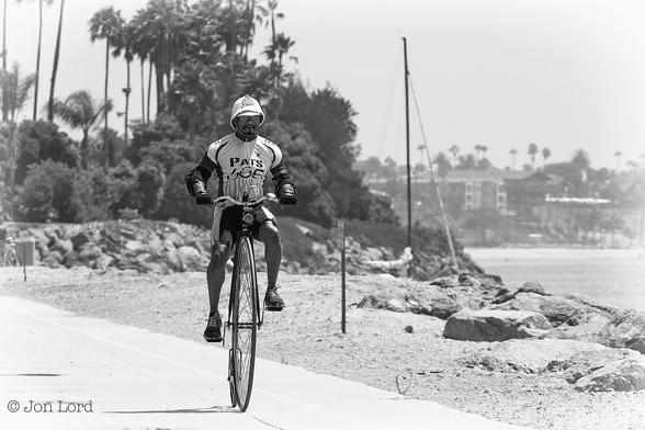 This is a black and white street photo of an eccentric looking man riding a Penny-Farthing. 
To the left of centre, facing and moving towards the camera is a man wearing a white pith helmet, aviator sunglasses and sports attire. He is riding a Penny-Farthing (sometimes called a High Wheel) bike. The bike has a large front wheel, perhaps a metre and a half with the pedals directly connected to the wheel (no chain or gears), a small set of handle bars with a small lantern and bell. Our rider sits on a saddle just behind the handle bars. Behind the large front wheel is a much smaller, maybe half a metre, wheel. The penny-farthing is on a concrete bike path that stretches diagonally across the lower left side of the frame. To the right is a short stretch of sand and then large rocks forming a harbour breakwater. In the distance to the left are some tall palm trees, while in the further distance to the right is a hazy view of some low-rise buildings above the coastline. 
The location: Long Beach, California. The tall palm trees are on "Island Grissom" an artificial island with oil wells hidden inside, the island named after the NASA astronaut Gus Grissom who was killed in the Apollo One fire (2014).