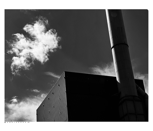 Schwarzweißfotografie: ein gestreifter blechverkleideter eckiger Industriekomplex mit Schornstein, dunkel gehaltene Fassade, starke Kontraste, grauer Himmel mit weißen Wolken, fotografiert mitten in der Stadt, in einem alten Fabrikgelände 