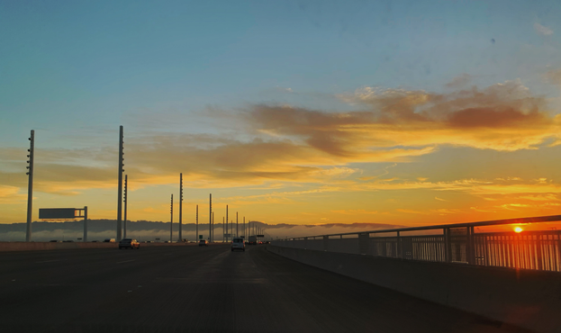 The sun setting over the road up to the San Francisco Bay Bridge. A bright orange-y sun can be seen peeking through the railing on the right side of the road. Yellow and gray clouds sit in front of a yellow and blue sky in the distance.