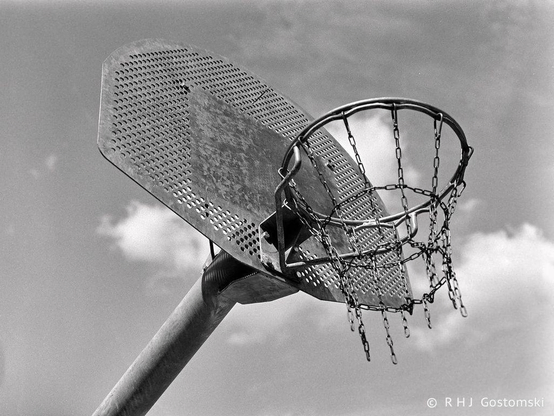 Black and white photo of a basketball hoop shot against a mostly clear sky with a few fluffy clouds