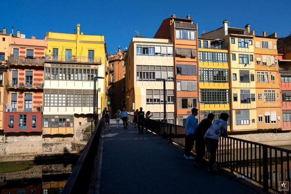 Color photo of pedestrians on a footbridge in deep shade, with brightly lit, colorful buildings in the background on the far side of the bridge. 