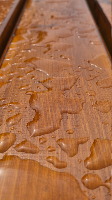 Nahaufnahme eines Holzbretts im Freien nach einem Regenguss. Es ist Teil einer Tischplatte und wurde der Länge nach fotografiert. Auf seiner lasierten Oberfläche haben sich Wasserlachen in verschiedenen Größen gebildet. Eine dieser Wasserlachen sieht aus wie ein Gespenst, das in einem typischen Gespenstertuch mit ausgestreckten Armen nach links schaut und in diese Richtung schwebt.

Close-up of a wooden plank outdoors after a downpour. It is part of a table top and was photographed lengthways. Puddles of water of various sizes have formed on its varnished surface. One of these pools of water looks like a ghost looking to the left in a typical ghostly scarf with outstretched arms and floating in that direction.