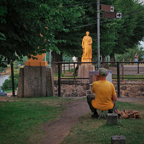 In the photo, the artist is sitting on a folding chair and painting on an easel. In front of him is a sculpture, which may have served as inspiration for the painting. Next to the artist is a bag that may contain his painting supplies. To the artist's left, another sculpture can be seen through the trees. The artist's T-shirt and the sculpture share a similar orange color.