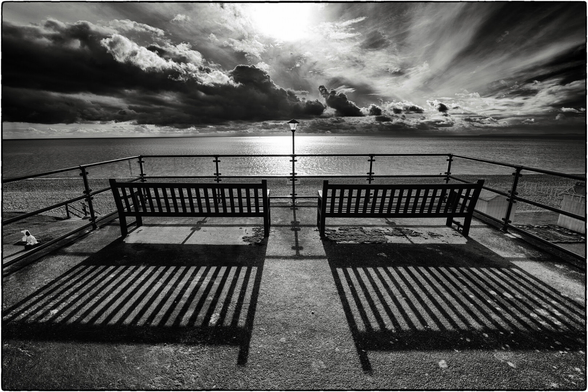 Schwarzweissfotografie einer dramatischen Meeresszene von einer Aussichtsplattform aus. Zwei leere Holzbänke stehen symmetrisch angeordnet mit Blick auf das Meer, getrennt durch eine klassische Straßenlaterne in der Mitte. Die Bänke und das Geländer werfen markante Schatten auf den gepflasterten Boden. Der Himmel dominiert das Bild mit dramatischen, dunklen Wolkenformationen, durch die Sonnenstrahlen brechen und das Wasser zum Glitzern bringen.