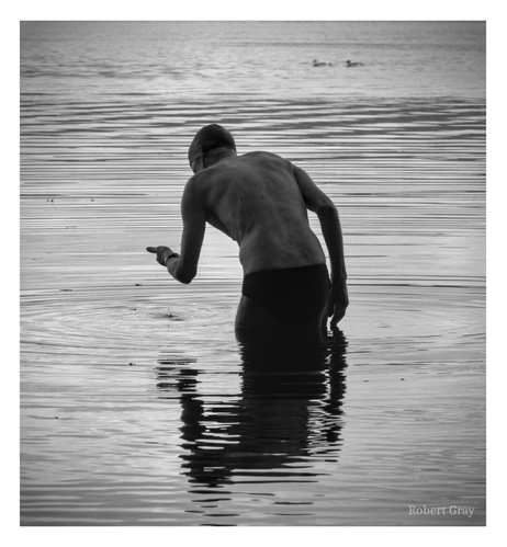 Black and white image of an old man, seen from behind, bending over and letting water drop from his hand.
