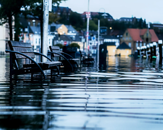 Die Sicht geht geradeso über die Wasseroberfläche hinweg. Vorne stehen mehrere Sitzbänke, die schon fast bis zu Sitzfläche in der Flut stehen, dahinter die erste Häuserreihe am Hafen von Flensburg. Die Flut reichte weit in die Stadt hinein und überflutete eine ganze Reihe Häuser. 