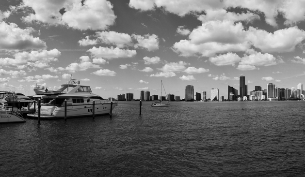 Black and white image of a marina with docked yachts and a distant city skyline under a sky filled with fluffy clouds, conveying a serene atmosphere.