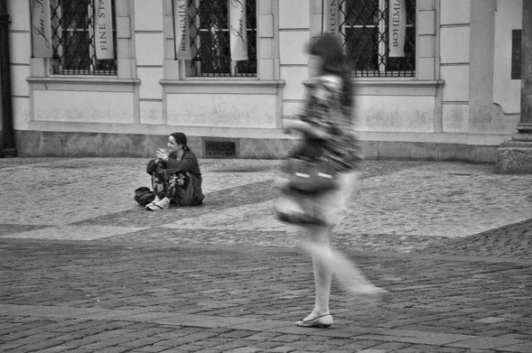 A blurred black and white photograph of a street scene. A person in motion is visible on the right, their body blurred from movement. Another person is sitting on the ground on the left. The ground is paved with cobblestones. In the background, there is a building with windows and banners.