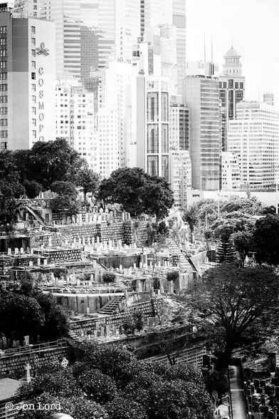 A Grungy Black And White Cityscape Photo In Portrait Format. In The Middle Ground Is A Hillside Cemetery With Numerous Tiers Cut Into The Hill That Are Lined With Headstones. Surrounding the Cemetery Are Mature Dark Trees That Almost Encircle The Hill. In The Background And Slightly Overexposed Is A White High-Rise, Very Dense Cityscape. The Cemetery On The Hillside Is The Happy Valley Muslim Cemetery, The Headstones At The Base Of The Image Are In The Saint Michael Catholic Cemetery. The High-Rise Buildings In The Background Are In Wan Chai, Hong Kong (2013).