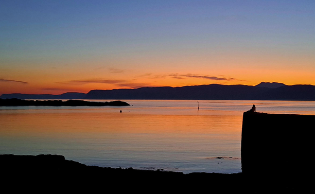 The sun has set, leaving a fiery orange sky fading upwards to celestial blue; this is reflected in an almost perfectly calm sea. All terrestrial features are reduced to solid black: the shore, a reef, the island of Mull across the Firth of Lorn. On the jetty above the beach, a figure, leaning back.on her arms, sits and watches the sky reveal its glories...