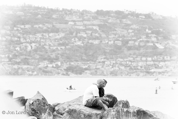This is a black and white street photo of a man and boy sitting on rocks by the sea. 
Stretching along the lower margin are a collection of large, medium grey, rocks that form a harbour breakwater. Below centre and ever so slightly to the right is a man and boy sitting on one of the larger and flatter rocks. The man is wearing a large and baggy white t-shirt with light grey shorts. The boy is wearing a black t-shirt and has his right arm around the mans shoulder. Both are looking downwards, perhaps at the water below the rocks. Behind the rocks is a bay with a few small boats and a few smaller buoys. In the background is the hazy and blurred view of rising terrain filled with multiple tiers of residential low rise buildings. Above is a narrow and sloping strip of hazy, milky white sky.
The location: Redondo Beach, California (2014).