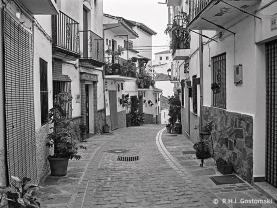 A black and white photo of a narrow cobbled street with white houses in Jubrique