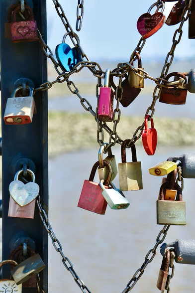 Verschiedene Vorhängeschlösser, teils in Herzform teils in normaler Form , hängen an einer Kette. Die Schlösser haben sind gelb, rot, blau, pink, silber und bronze. Im Hintergrund ist verschwommen Wasser zu erkennen. 