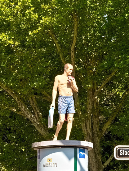 On an advertising column stands the lifelike sculpture of a man. He is only wearing blue shorts, his upper body is bare. In his right hand he carries flippers, his left hand holds a towel, which he has thrown loosely over his shoulder. He is looking slightly to the left and appears relaxed. The sun is shining on him.
These types of sculptures can be found all over Düsseldorf.