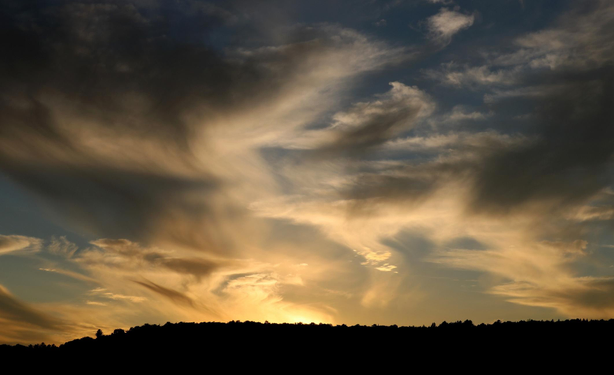 Photograph of a sunset in a cloudy sky, with a yellow light coming from the sun just getting under the horizon, and weird grey and white clouds, as if someone had dragged a paintbrush over oil paint in the sky.