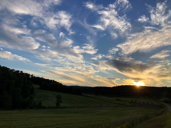 Photograph of a sunset in a cloudy blue sky, over a rural landscape of hilly green fields with bales of hay and forest in the distance.