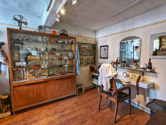 A well-preserved, vintage hairdresser's shop interior, likely from the mid-20th century. The room has light-coloured walls with an arched decorative trim, and a rustic wooden floor. On the left, a large, dark wood and glass display cabinet is filled with various old-fashioned hairdressing tools, products, and equipment. An old electric fan sits on top of the cabinet. In the centre is a classic, dark wooden barber's chair with a white cloth draped over it. To the right, a white ceramic sink and a large mirror are mounted on the wall, with an array of shaving brushes, bottles, and other grooming implements displayed on the shelf. The space has a nostalgic feel, showing the tools of the trade from a bygone era.
