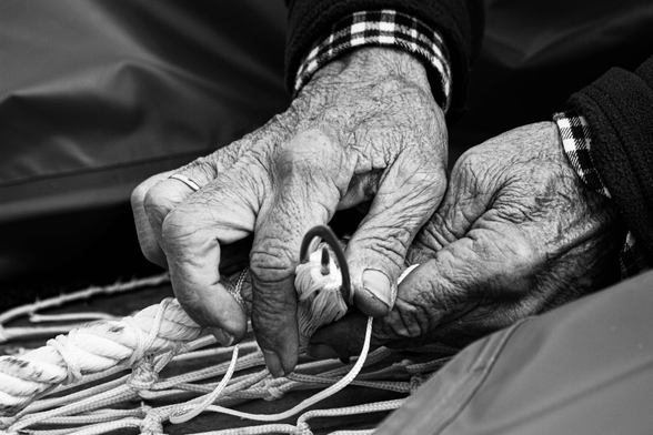 A black and white photograph of a fisherman's hands mending a net. You can see the net in the lower left corner, and in the center and upper right corners, two weathered and wrinkled hands, overlapping each other. The right hand above holds a small, oval instrument with a pin between its index finger and thumb, while the lower hand holds the net.