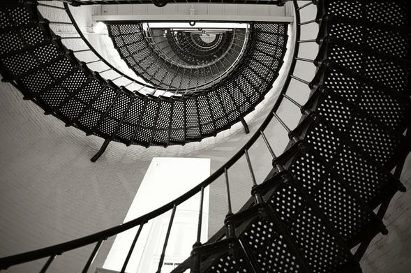 A black-and-white image of a lighthouse's spiral staircase, viewed from below. It creates a dizzying pattern of concentric circles ascending upward.