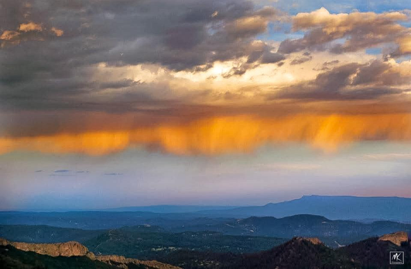 Color photo of a western US landscape taken looking towards the east from the slopes of Colorado’s Spanish Peaks, with the setting sun lighting up the virga under a dying summer rainstorm. 