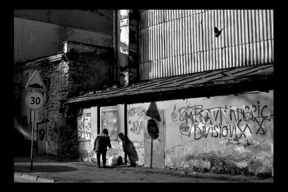 A gritty black and white urban scene: a person in a hooded coat bends near a wall covered in graffiti and posters, casting a long shadow. Above, a bird is caught mid-flight against corrugated metal. The decaying brick and plaster add texture, while a road sign marks a speed limit of 30.